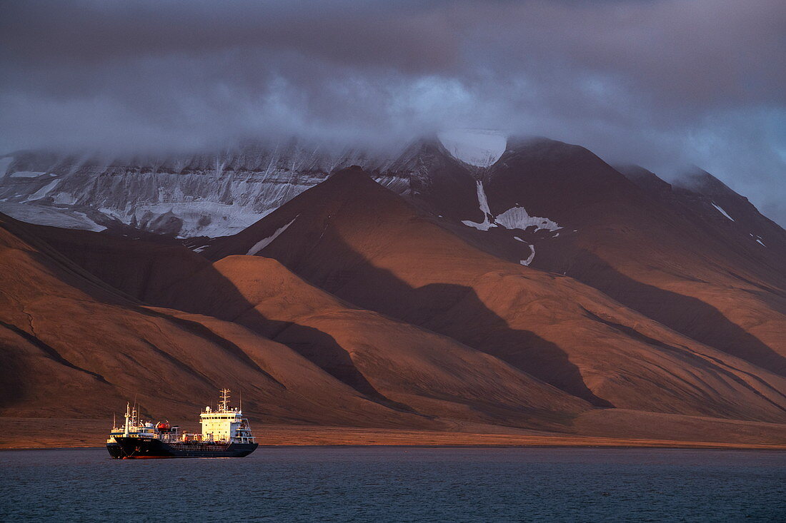 Die späte Abendsonne malt eine besondere Szene, als ein Schiff an kargen, rötlichen Bergen vorbeifährt, Longyearbyen, Spitzbergen, Norwegen, Europa