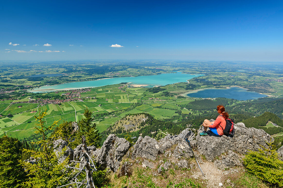 Woman hiking sits on rock and looks at Forggensee and Bannwaldsee, Tegelberg, Ammergau Alps, Swabia, Bavaria, Germany