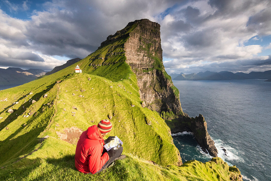 Wanderer betrachtet die Landkarte auf einer Klippe über dem Ozean in Richtung Kallur Leuchtturm, Insel Kalsoy, Färöer-Inseln, Dänemark