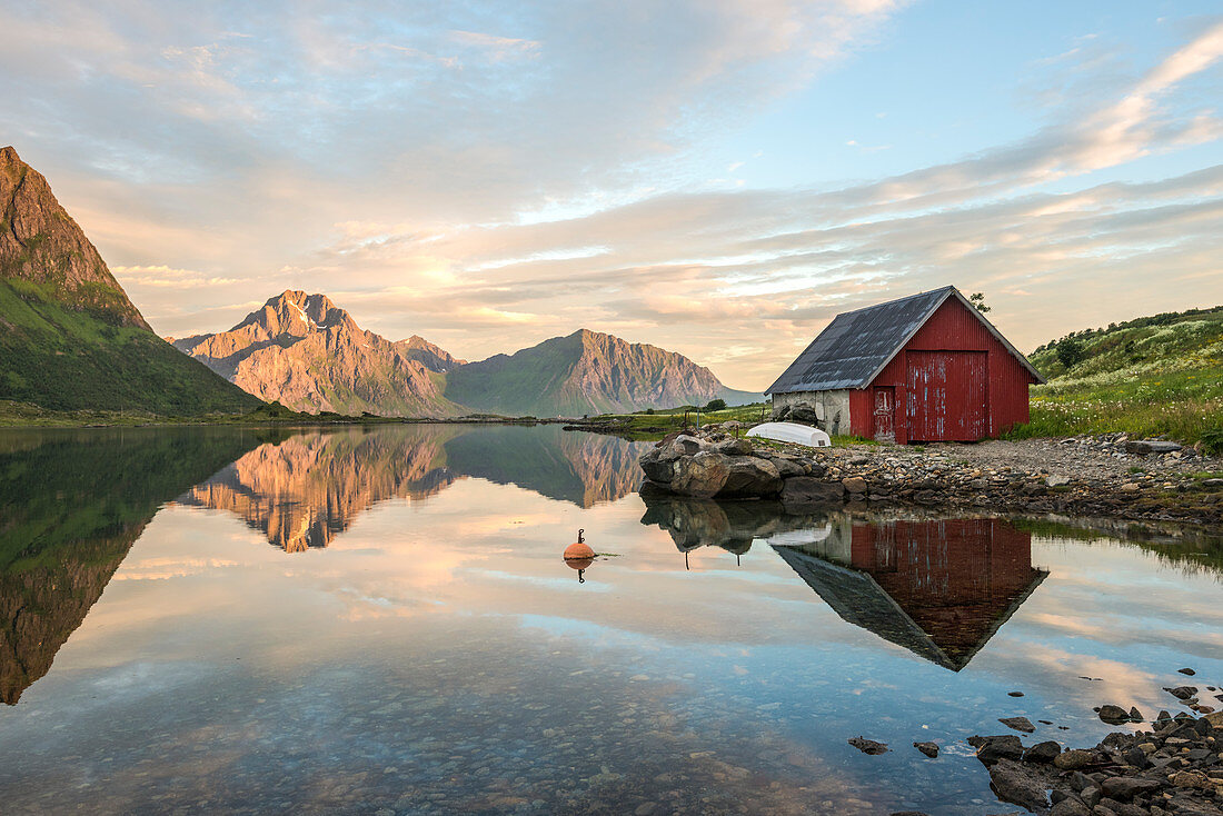 Typical rorbu and peaks reflected in the clear sea during midnight sun, Vengeren, Vagpollen, Lofoten Islands, Norway, Europe