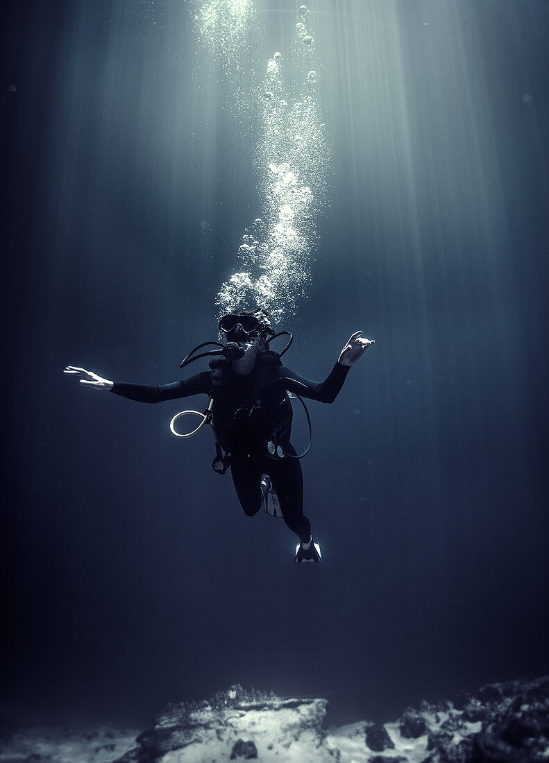 Underwater view of diver wearing wetsuit, diving goggles and oxygen cylinder, air bubbles rising.