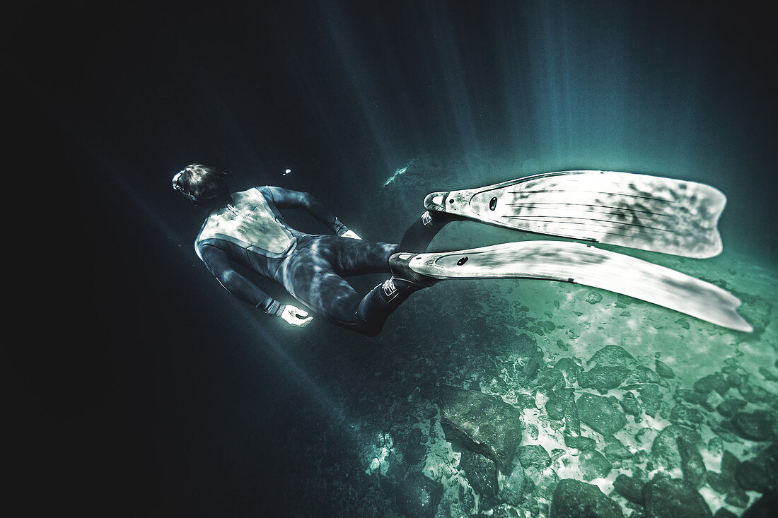 High angle underwater view of diver wearing wet suit and flippers, sunlight filtering through from above.