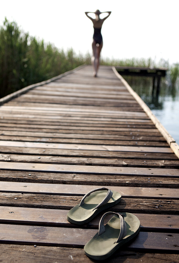 Unscharfe Rückenansicht einer Frau im Badeanzug, die über Bootssteg läuft, im Vordergrund ein Paar Flip-Flops