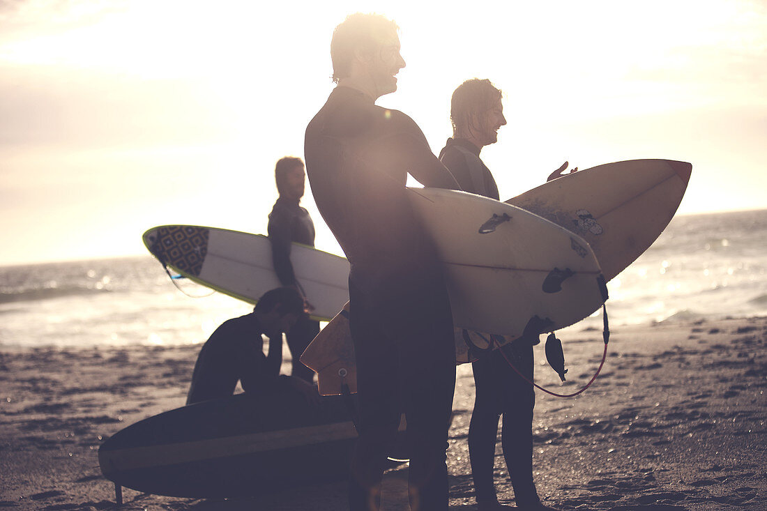 Four men wearing wetsuits standing on a sandy beach, carrying surfboards.