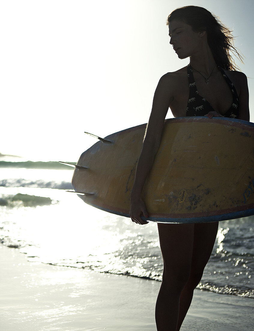 Woman standing on a sandy beach by the ocean, holding a surfboard.