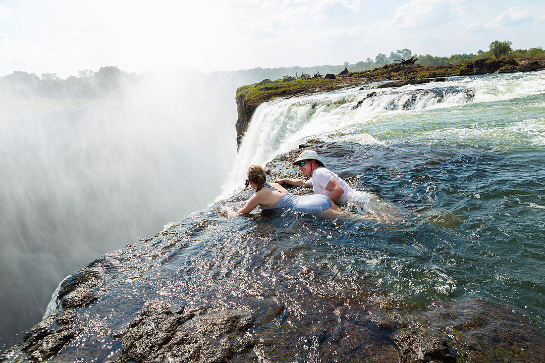 Mann und junge Frau im Wasser des Devils Pool am Rande der Victoriafälle, aufsteigender Nebel über dem Wasserfall