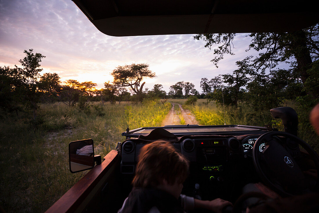 Safari-Jeep fährt mit eingeschalteten Scheinwerfern nach Sonnenuntergang auf einem Feldweg entlang