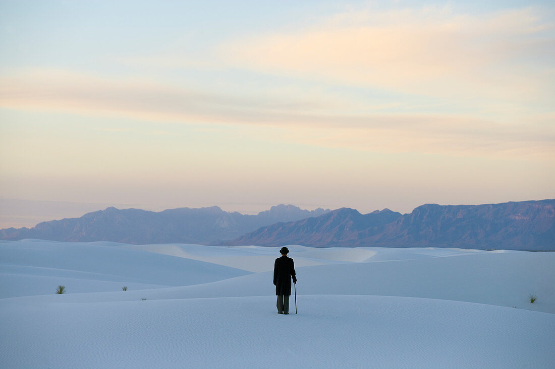 Man in a black coat and suit, a bowler hat and umbrella, in a white desert wilderness of white sand.