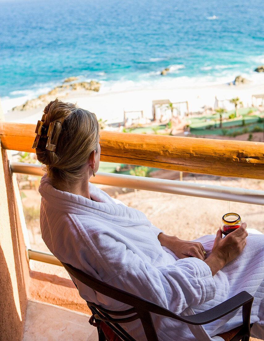 Frau sitzt mit einem Getränk auf einem Hotelbalkon mit Blick auf das Meer und den weißen Sandstrand