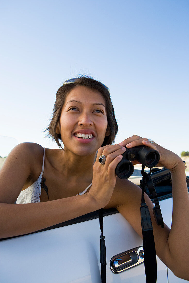 Native American woman in sun dress driving a white convertible sports car looking through binoculars