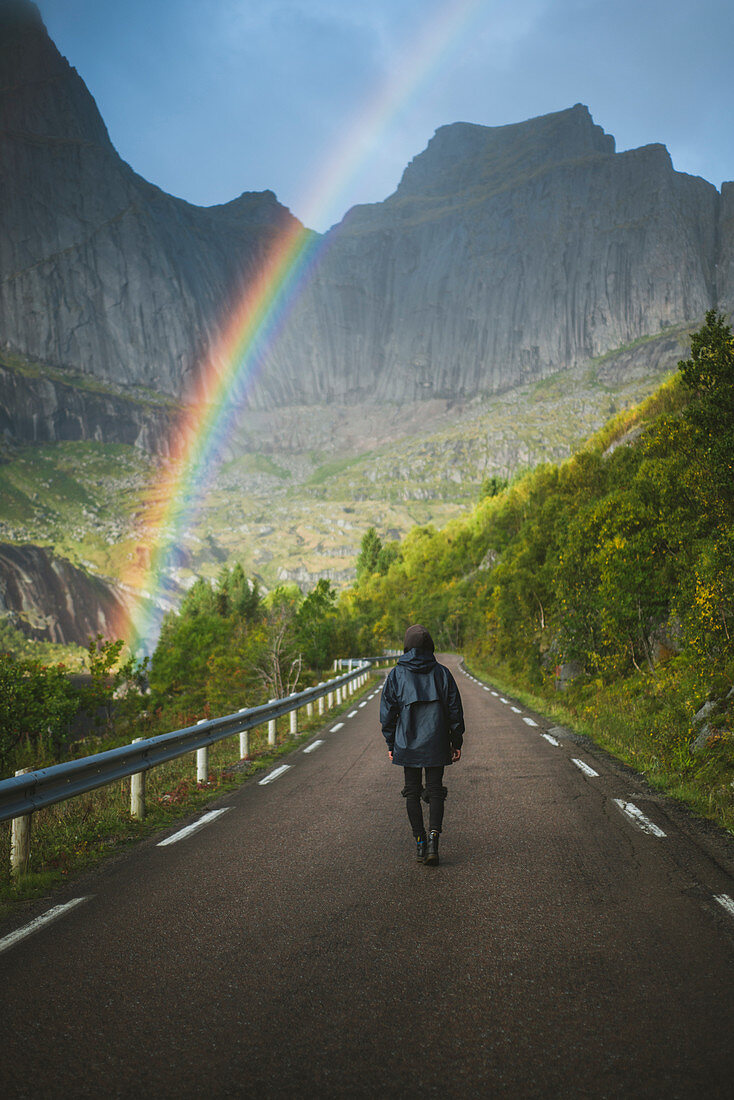 Norwegen, Lofoten, Mann auf der Straße mit Bergen und Regenbogen im Hintergrund