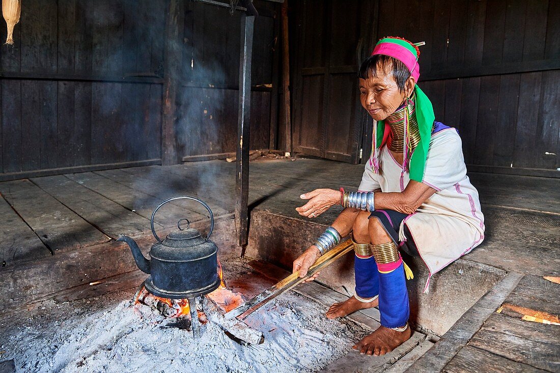 Kayan Lahwi Frau mit Messinghalsringen und traditioneller Kleidung, kocht Wasser in ihrem Haus, um zu ihrem Lieblingsgetränk einen aus fermentiertem Sorghum (links im Glas) destillierten Schnaps hinzuzufügen. Pan Pet Region, Bundesstaat Kayah, Myanmar.