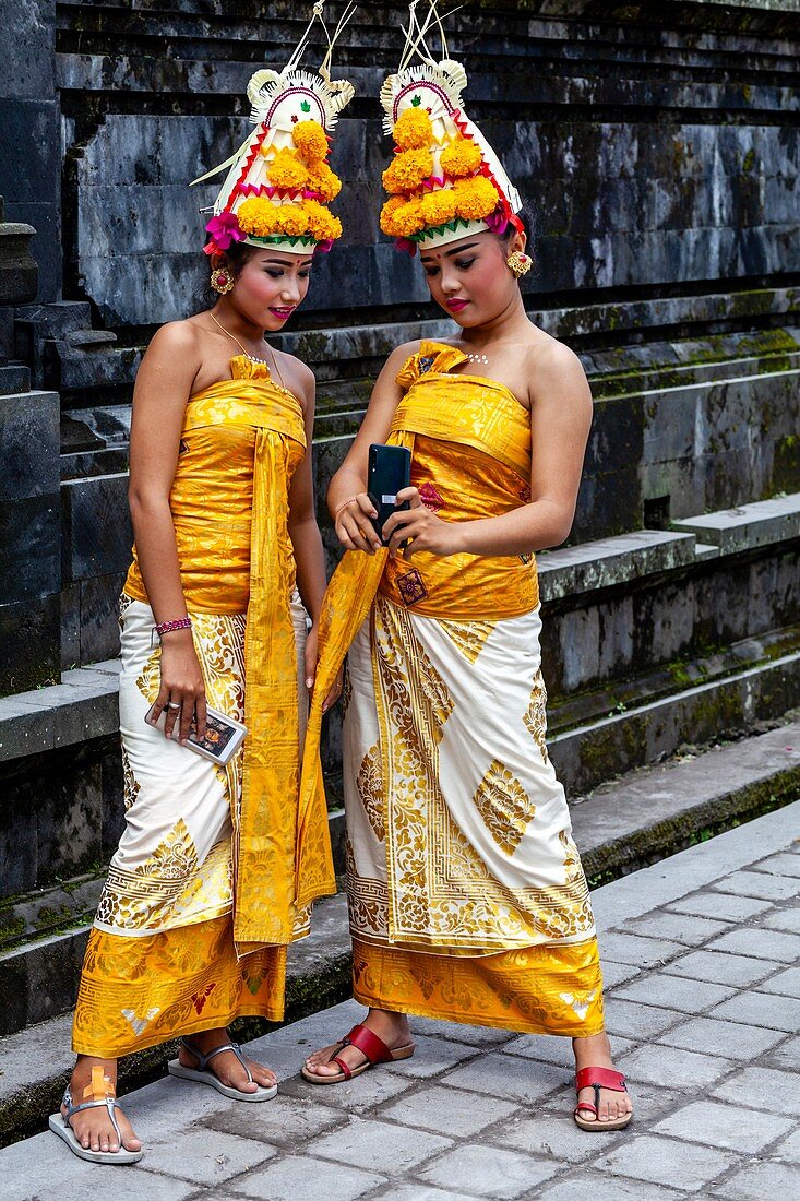 Zwei junge balinesische Hindu-Frauen mit Blick auf ein Mobiltelefon (Handy) bei der Batara-Turun-Kabeh-Zeremonie, Besakih-Tempel, Bali, Indonesien