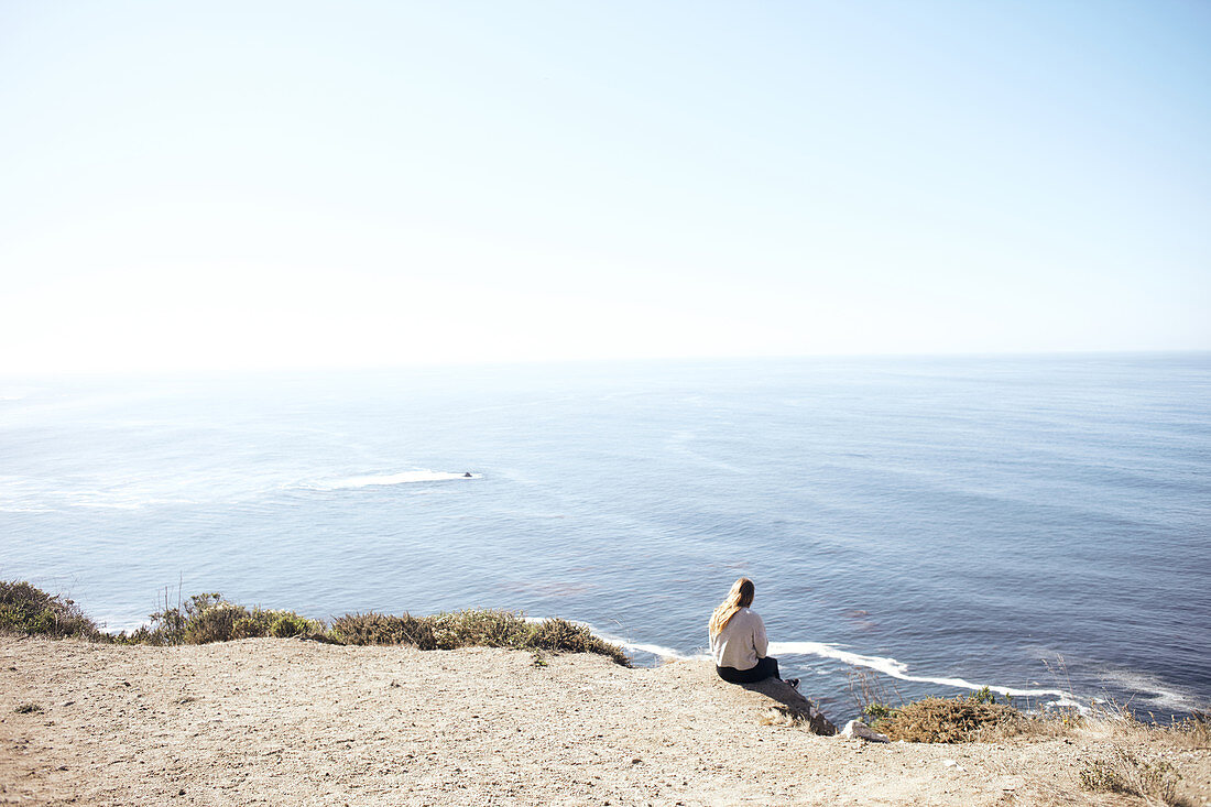 Young woman sits on a cliff and looks out over the Pacific Ocean on the edge of Highway 1, Big Sur State Park, California, USA.
