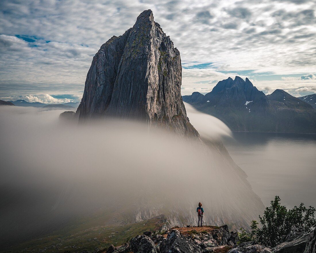 HIKER AT THE FOOT OF MOUNT SEGLA SURROUNDED BY CLOUDS PLUNGING INTO THE FJORD, FJORDGARD, SENJA, NORWAY
