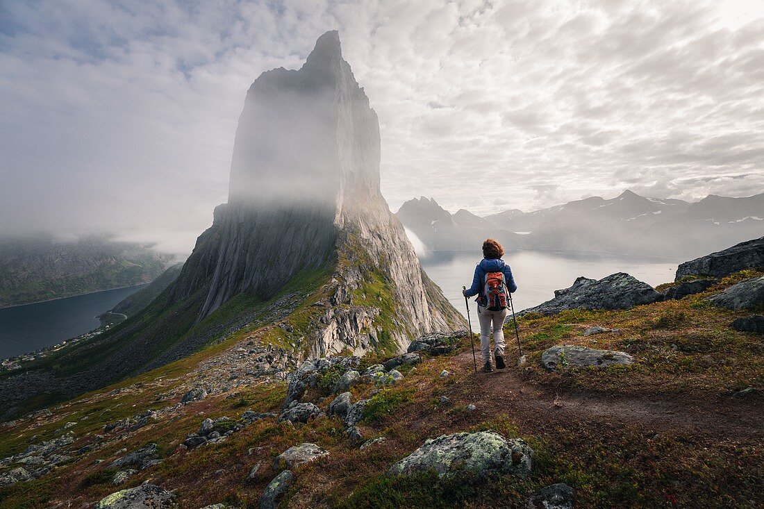 HIKER APPROACHING THE CLIFF FACES OF MOUNT SEGLA PARTIALLY HIDDEN BY THE CLOUDS, FJORDGARD, SENJA, NORWAY