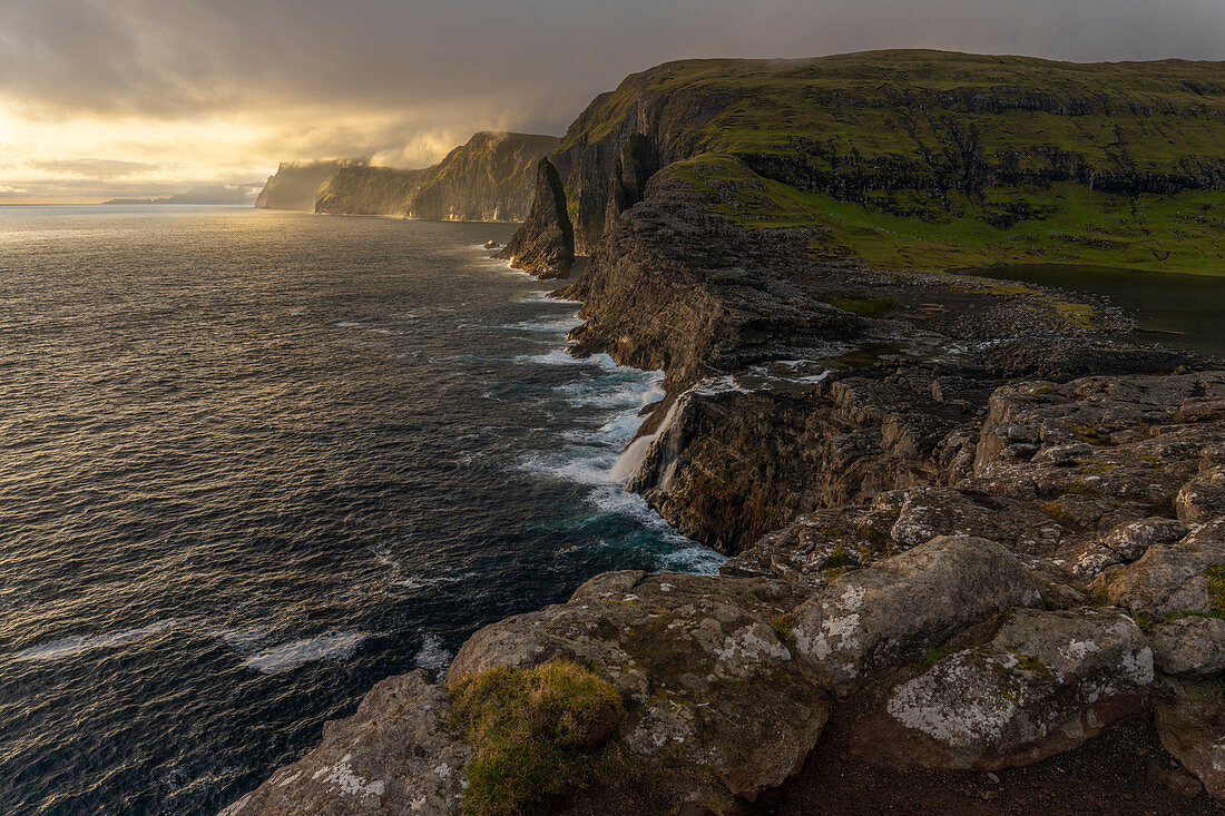 Steep coast in the west of the island of Vágar with the waterfall Bøsdalafossur and the rock needle Geitaskoradrangur near the largest lake in the Faroe Islands, Leitisvatn, Faroe Islands
