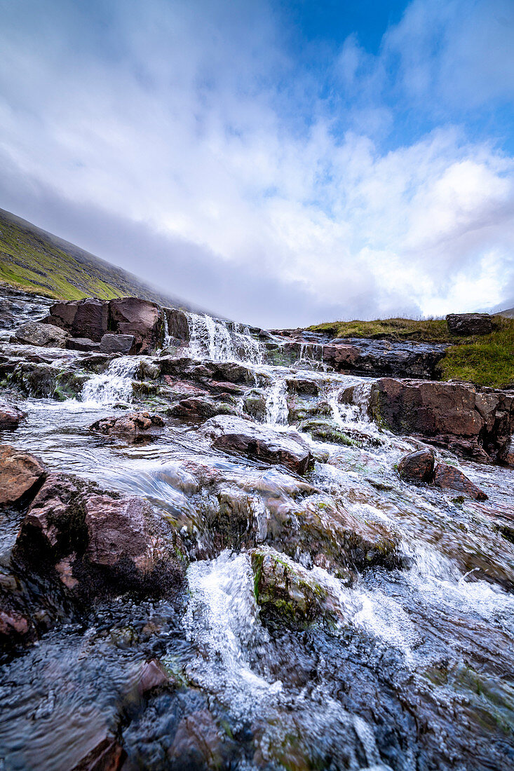 Bachlauf und kleiner Wasserfall auf der Insel Streymoy mit wolkigem Himmel, Färöer Inseln