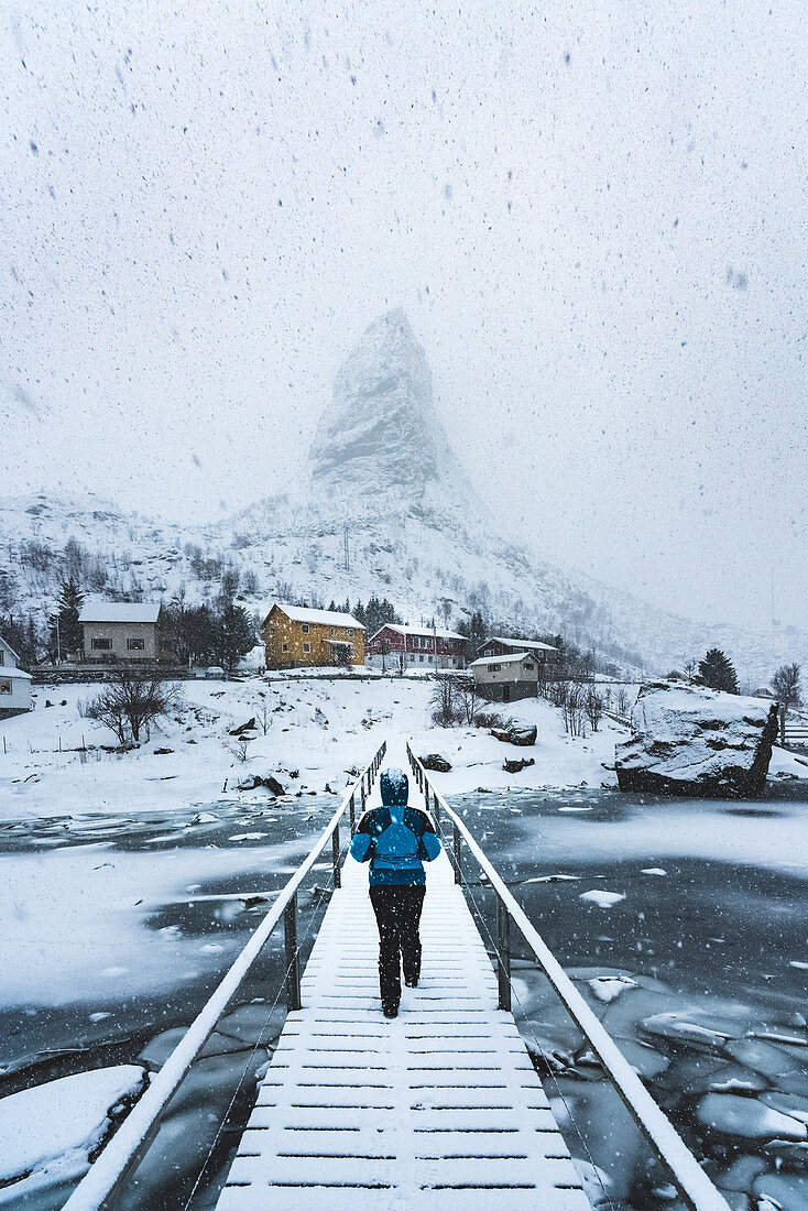 Frau geht über einer Holzbrücke im Schnee mit Blick Richtung Hammarskaftet, Reine, Nordland, Nordnorwegen, Norwegen