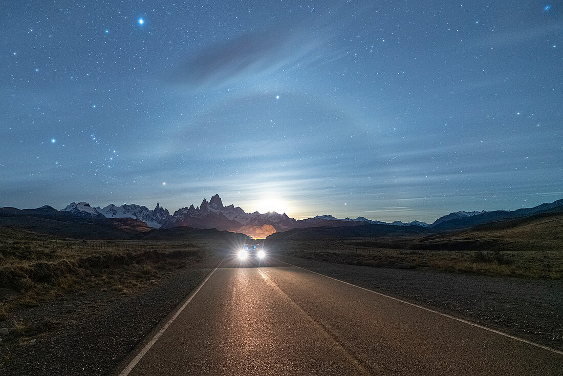 Car driving on the road leading to El Chalten, with Fitz Roy range and moonlight in the background at night. El Chalten, Santa Cruz province, Argentina.