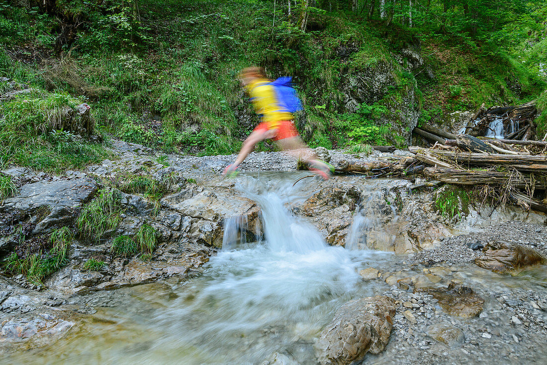 Woman hiking jumps over creek, Kraxenbachtal, Sonntagshorn, Chiemgau Alps, Chiemgau, Upper Bavaria, Bavaria, Germany