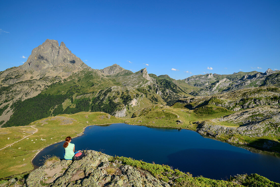 Frau beim Wandern sitzt auf Felssporn und blickt auf Lac Roumassot und Pic du Midi d´Ossau, Lac Roumassot, Nationalpark Pyrenäen, Pyrénées-Atlantiques, Pyrenäen, Frankreich