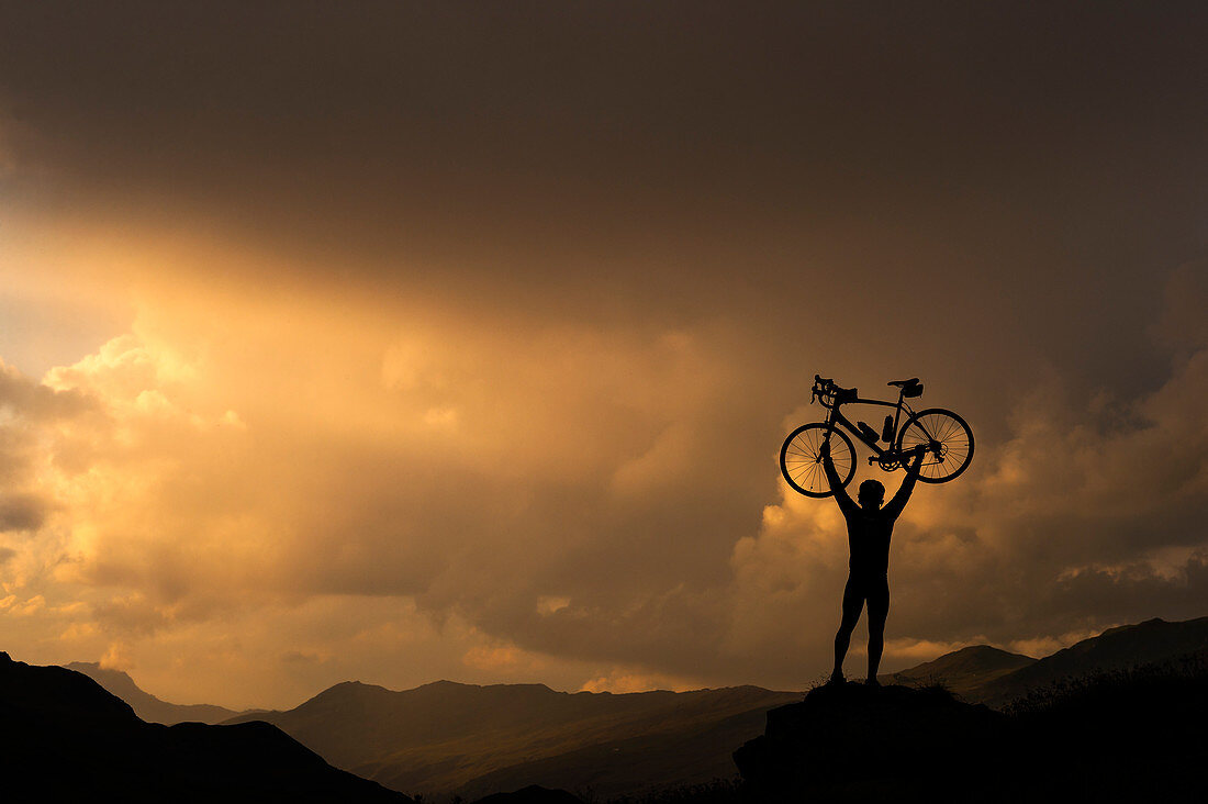 Radfahrer, der Fahrrad gegen Sonnenuntergang auf Hügel hebt