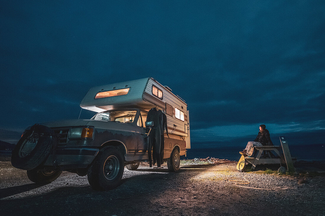 Woman sitting on picnic bench next to camper-van parked near Jordan River, British Columbia, Canada at night.