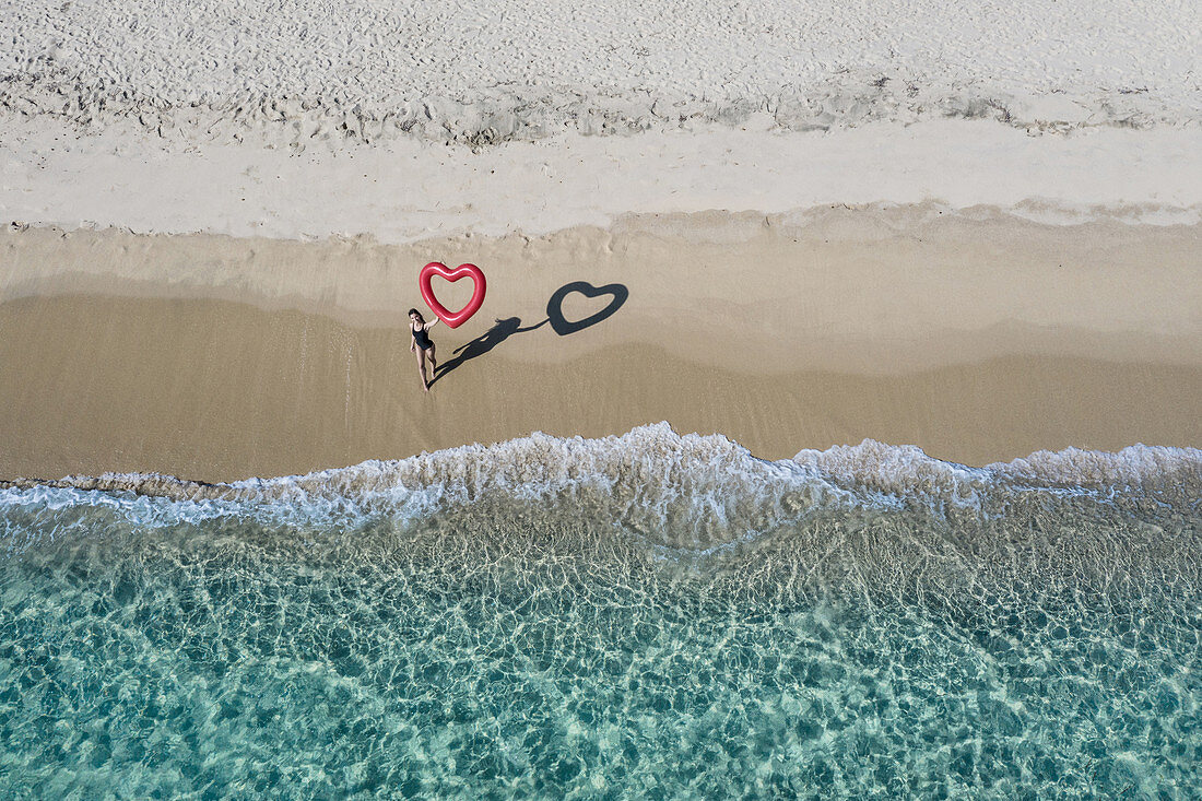 Luftaufnahme der jungen Frau im Badeanzug, die auf einem Sandstrand steht und roten herzförmigen Ballon hält.