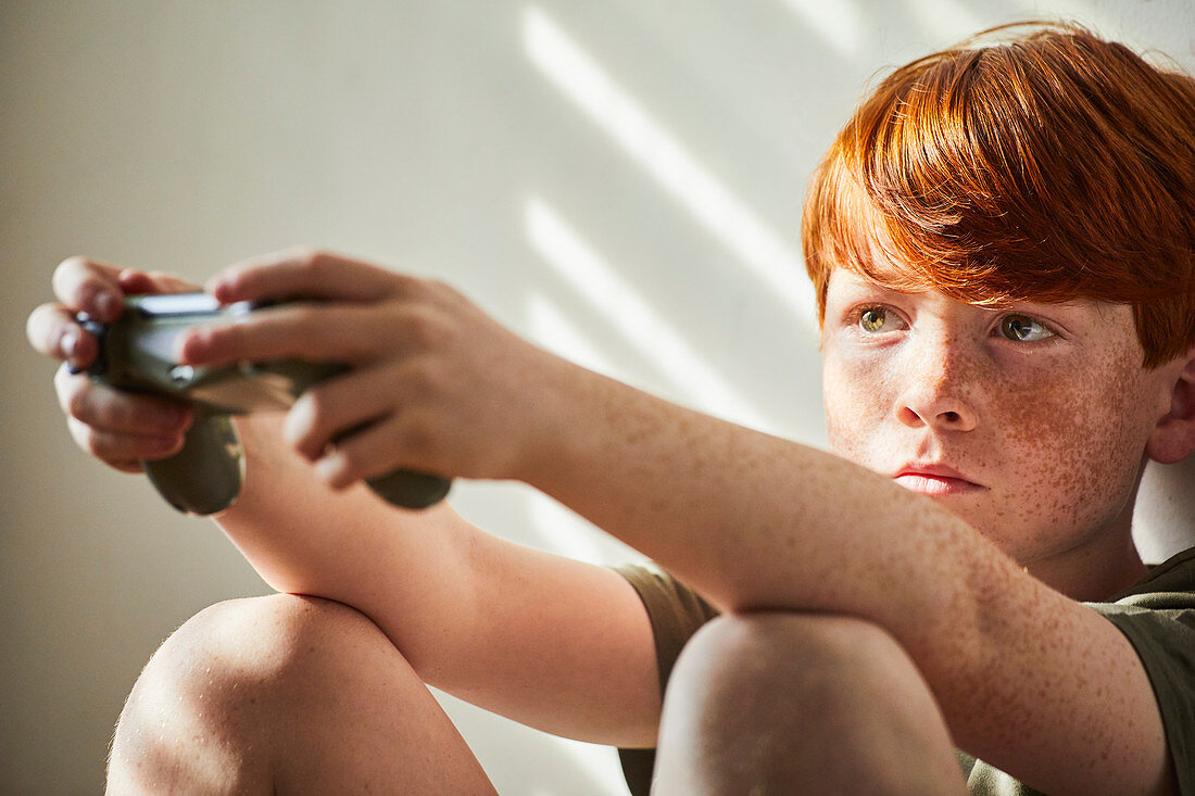 Junge mit den roten Haaren, die auf Boden in sonnigem Raum sitzen und Spielkonsolensteuerung halten.
