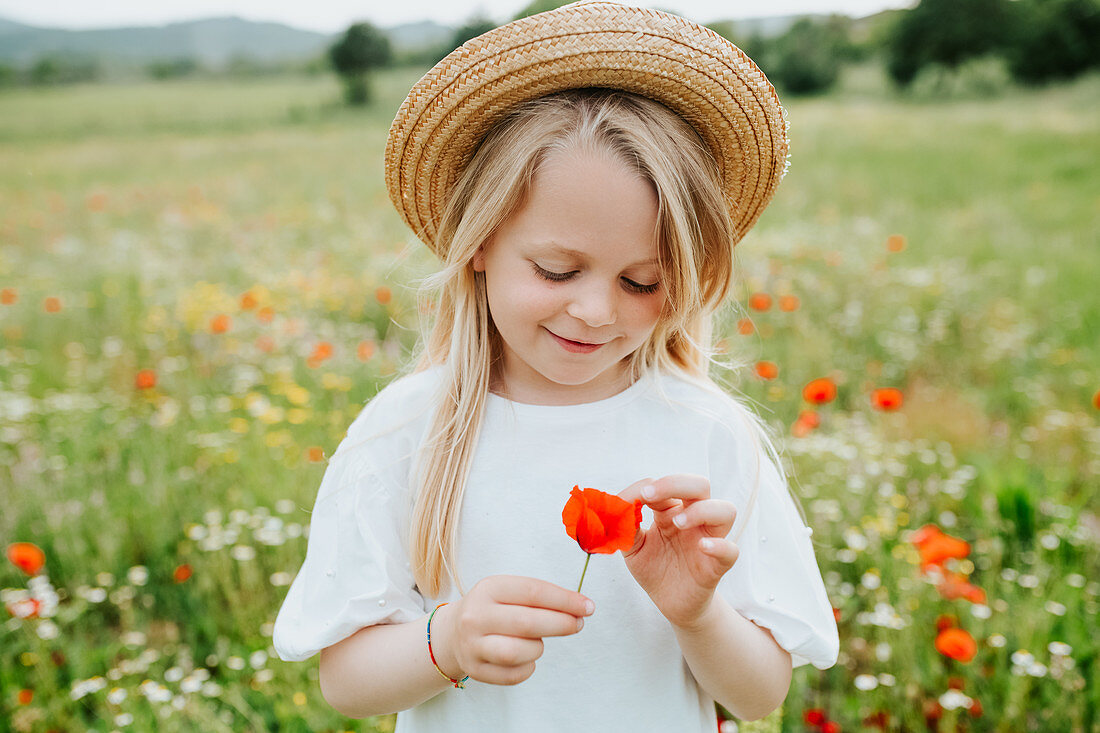 Portrait of young girl holding a poppy, standing in a meadow of wild flowers.