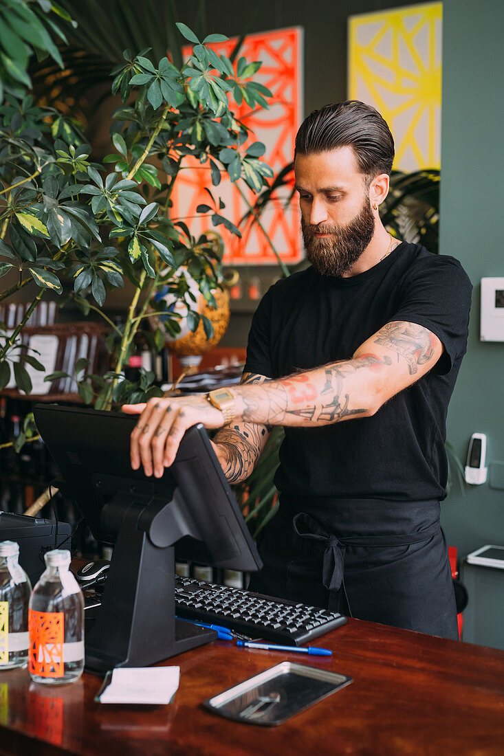 Tätowierter Barkeeper mit Bart an der Theke stehend benutzt eine computergesteuerte Kasse