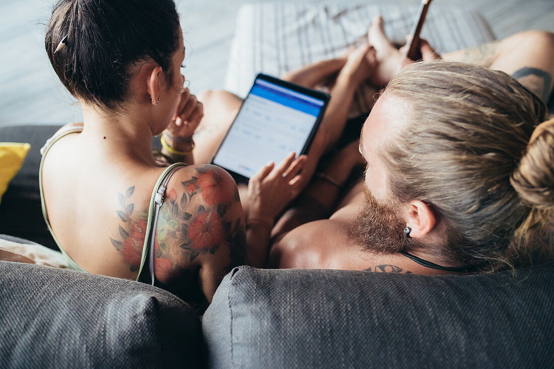 Bearded tattooed man with long brunette hair and woman with long brown hair sitting on a sofa, looking at digital tablet.