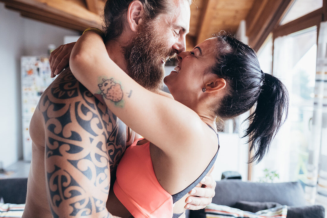 Bearded tattooed man with long brunette hair and woman with long brown hair standing indoors, hugging and kissing.