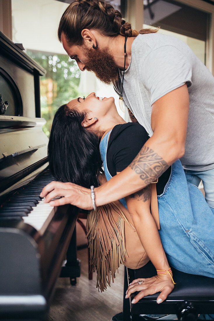 Bearded tattooed man with long brunette hair leaning over smiling woman, playing piano.