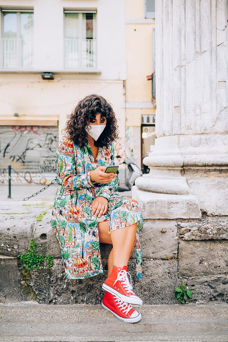 Young woman wearing face mask during Corona virus, sitting outdoors, checking mobile phone.