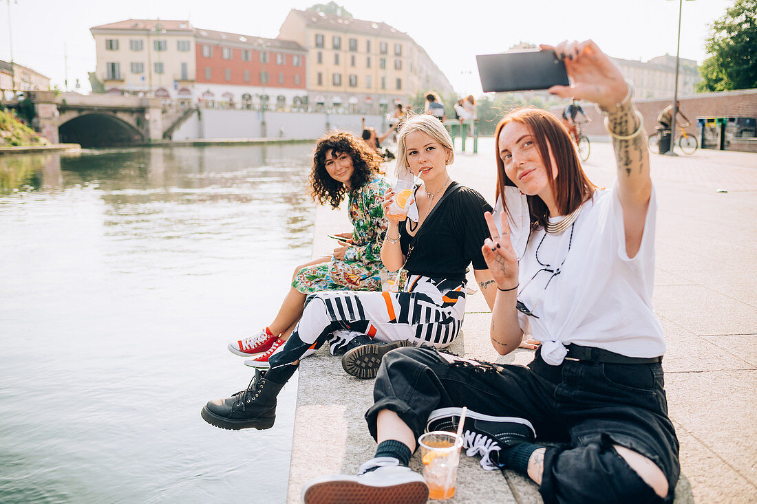 Three young women sitting on a riverbank, taking selfie.