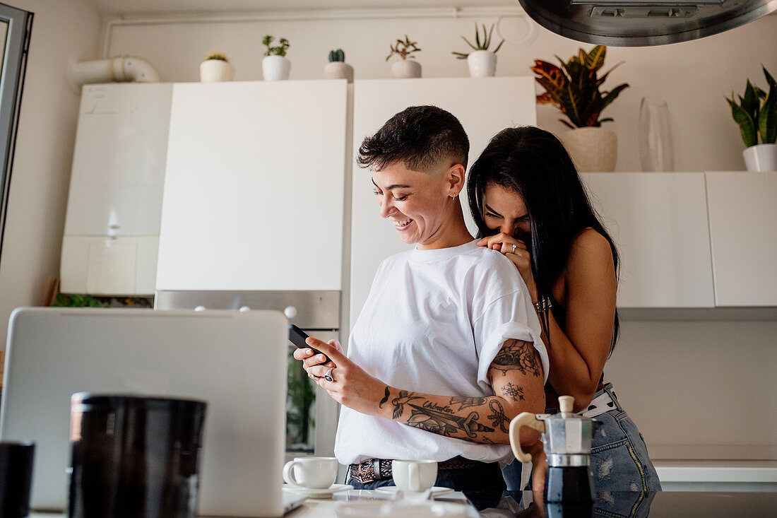 Young lesbian couple standing in kitchen, looking at mobile phone.