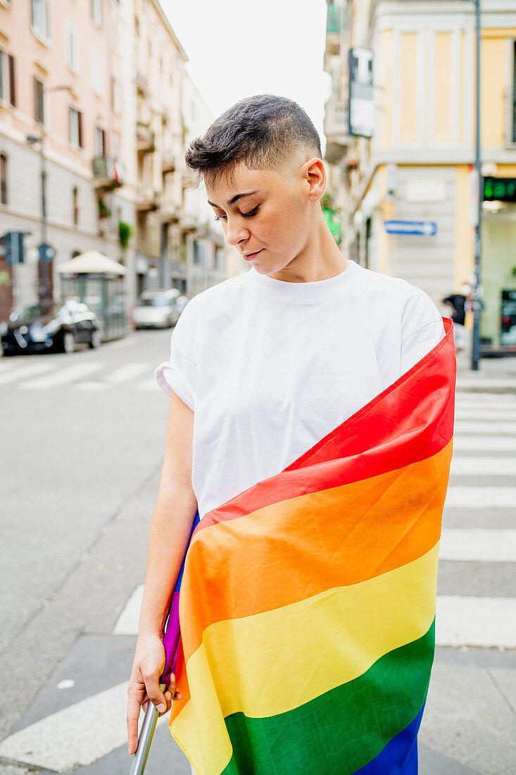 Young lesbian woman standing on a street, wrapped in rainbow flag.