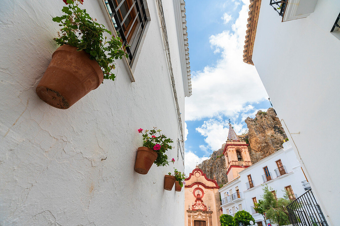Hängende Blumentöpfe an Gebäuden mit Maria De La Mesa-Kirche (Iglesia de Santa Maria de la Mesa) im Hintergrund, Zahara de la Sierra, Provinz Cadiz, Andalusien, Spanien