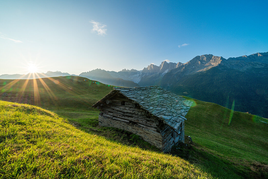 Steinhütte in grünen Weiden beleuchtet durch warmen Sonnenstich im Morgengrauen, Tombal, Soglio, Val Bregaglia, Kanton Graubunden, Schweiz