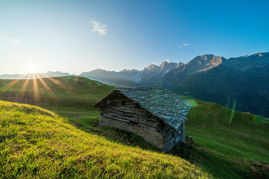 Stone hut in green pastures lit by warm sunburst at dawn, Tombal, Soglio, Val Bregaglia, canton of Graubunden, Switzerland