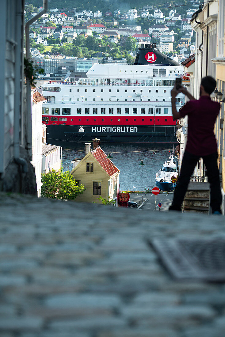 Rear view of man photographing the Hurtigruten cruise ship with smartphone, Bergen, Hordaland County, Norway