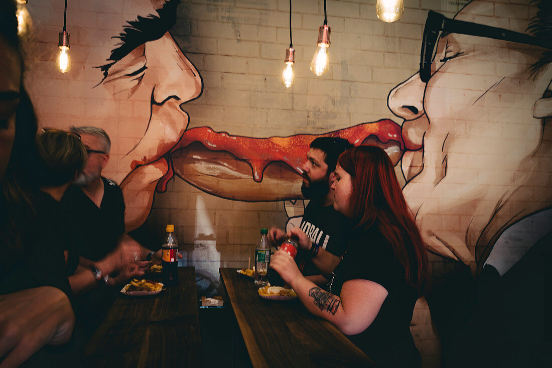 Menschen, die Currywurst essen, unter dem berühmten Bild von Erich Honecker und Leonid Brezenev, die sich küssen. Bezirk Mitte, BErlin, GErmany