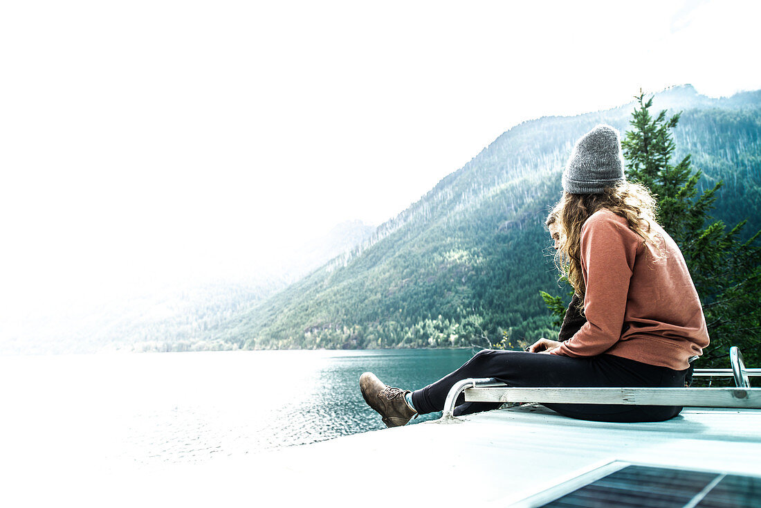 Frau sitzt auf dem Dach eines Wohnmobils mit Blick auf den See, Cathedral Grove, British Columbia, Kanada