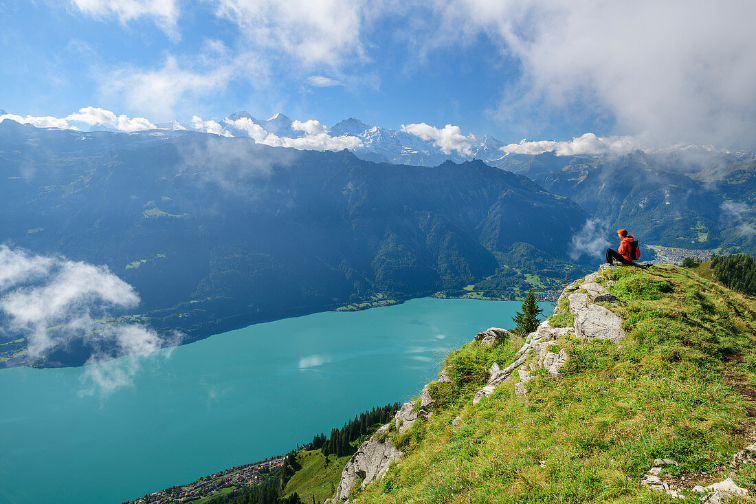 Woman while hiking sits on meadow ledge and looks out over Lake Brienz and the Bernese Alps in the background, from Augstmatthorn, Emmental Alps, Bern, Switzerland