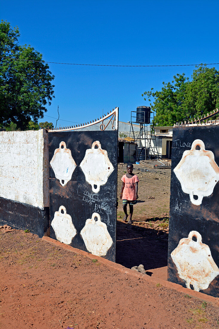 Gambia; Central River Region; Kuntaur; on the main street; Wall with half-open entrance gate to private property; Girl stands behind the gate and looks out at the street