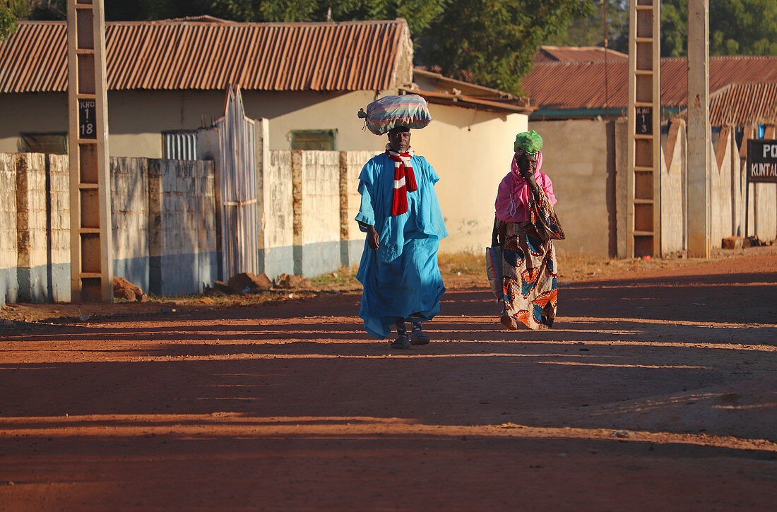 Gambia; Central River Region; Main street of Kuntaur; Married couple with luggage on their heads; Street scene in the morning sun;