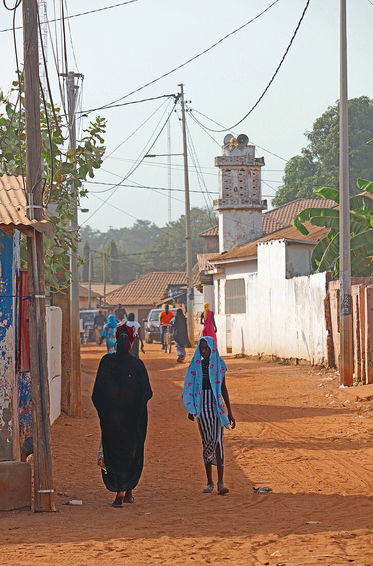 Gambia; Hauptstadtregion Banjul; Straßenszene in Bakau, in der Nähe des Kachikally Krokodilpools; im Vordergrund eine Frau im schwarzen Kleid und ein Mädchen mit türkisfarbenem Kopftuch;