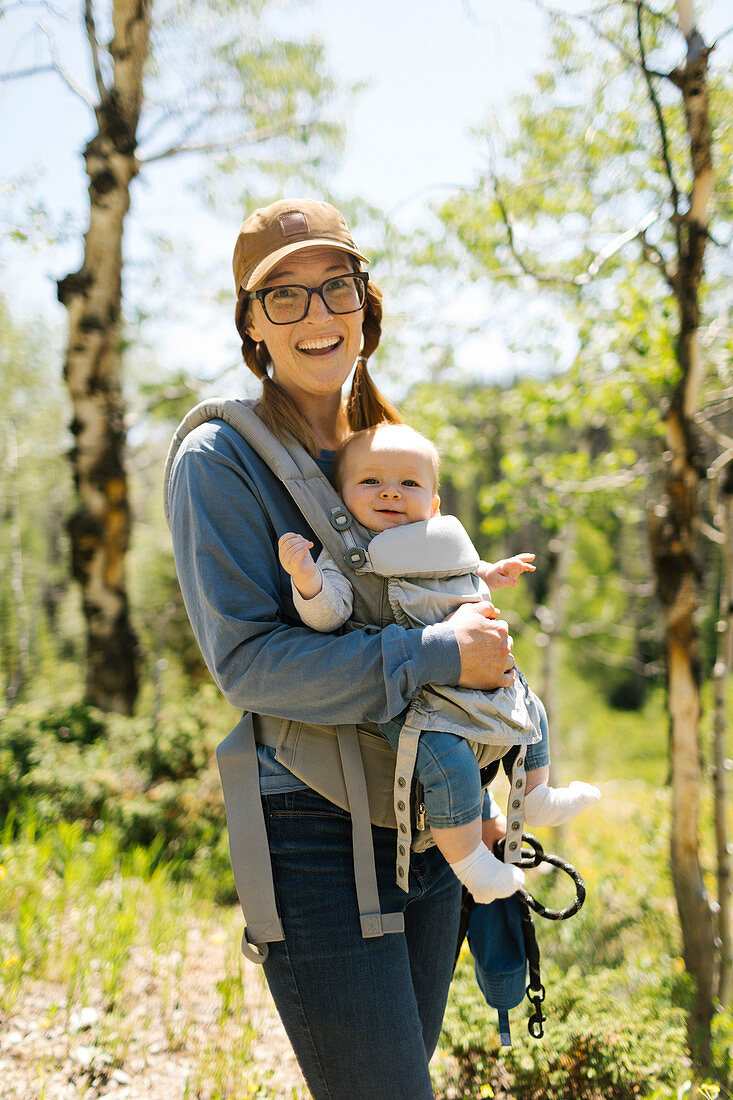Porträt einer lächelnden Frau mit dem kleinen Sohn in der Babytrage im Wald, Uinta-Nationalpark, Utah, USA